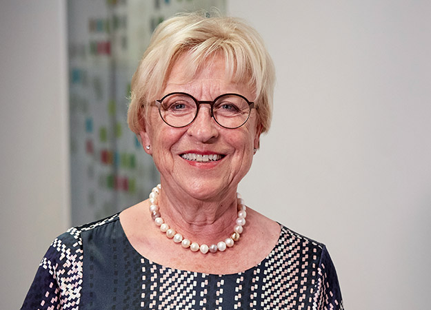 Smiling elderly woman wearing glasses, a patterned blouse, and a pearl necklace, standing in a bright indoor space.