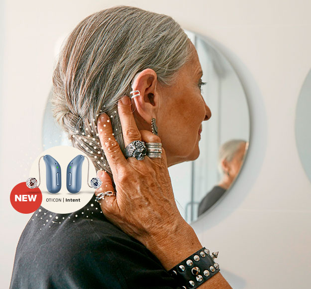 Person with grey hair adjusts their hearing aid while looking in a mirror. Text reads: New Oticon hearing devices.
