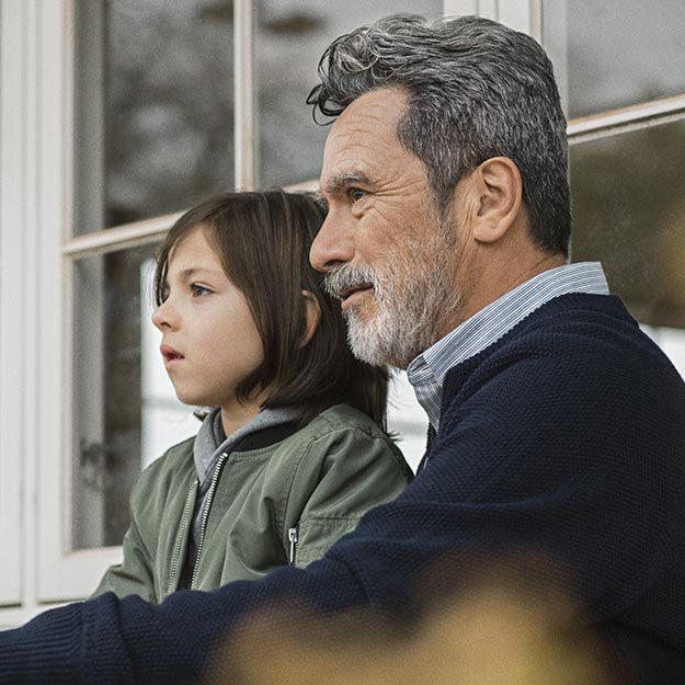 A gray-haired man, wearing a navy sweater and striped shirt, sits beside a child in a green jacket. They are outdoors near a window, suggesting a calm, home-like environment. No text visible.
