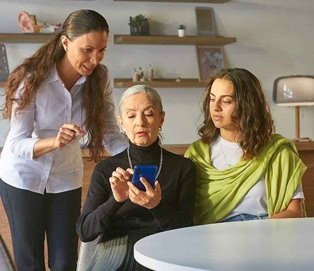 Three people are seated in a modern hearing clinic. A professional in a white shirt leans in, guiding an older individual interacting with a blue smartphone, while another person observes. Shelves in the background display hearing care materials, including ear models.