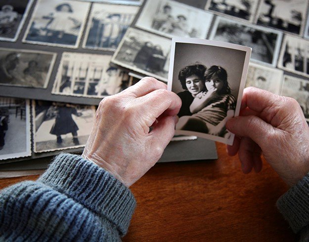 Hands of an older adult holding a vintage photo with blurred faces, surrounded by an assortment of black-and-white photographs on a wooden table.
