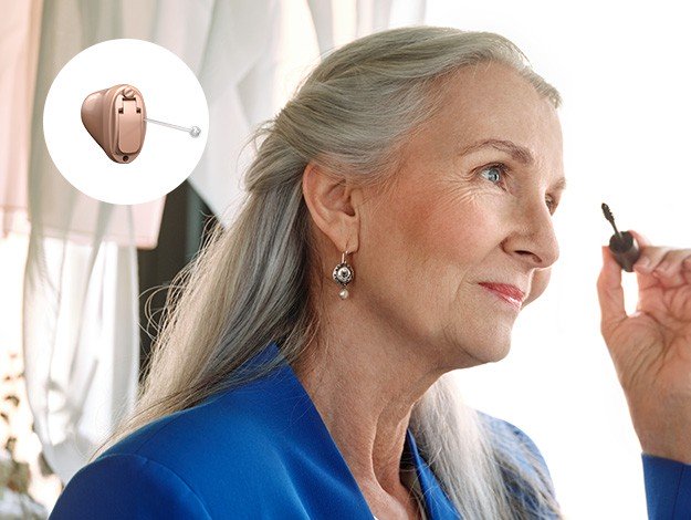 Elderly woman with long grey hair wearing a blue top inserts a small brown hearing aid into her ear. Inset image shows a close-up of the hearing aid. Background shows curtains.