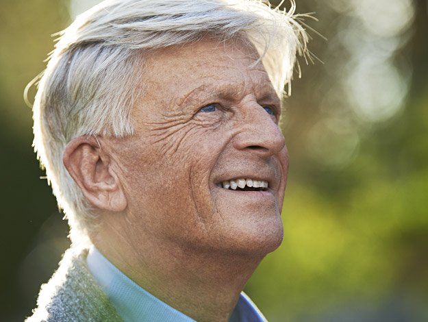 An older man with silver hair stands outdoors in a sunlit setting, wearing a blue collared shirt and sweater. His ear is visible, potentially emphasizing hearing care or hearing aids. No text is present.