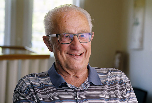 An elderly man with gray hair and glasses is smiling, wearing a striped polo shirt, seated indoors with blurred background elements of a railing and wall decor.