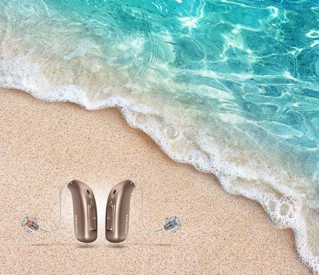 Two Oticon hearing aids are positioned on sandy beach terrain near the edge of clear turquoise ocean waves, symbolizing advanced hearing care in a serene, natural setting.