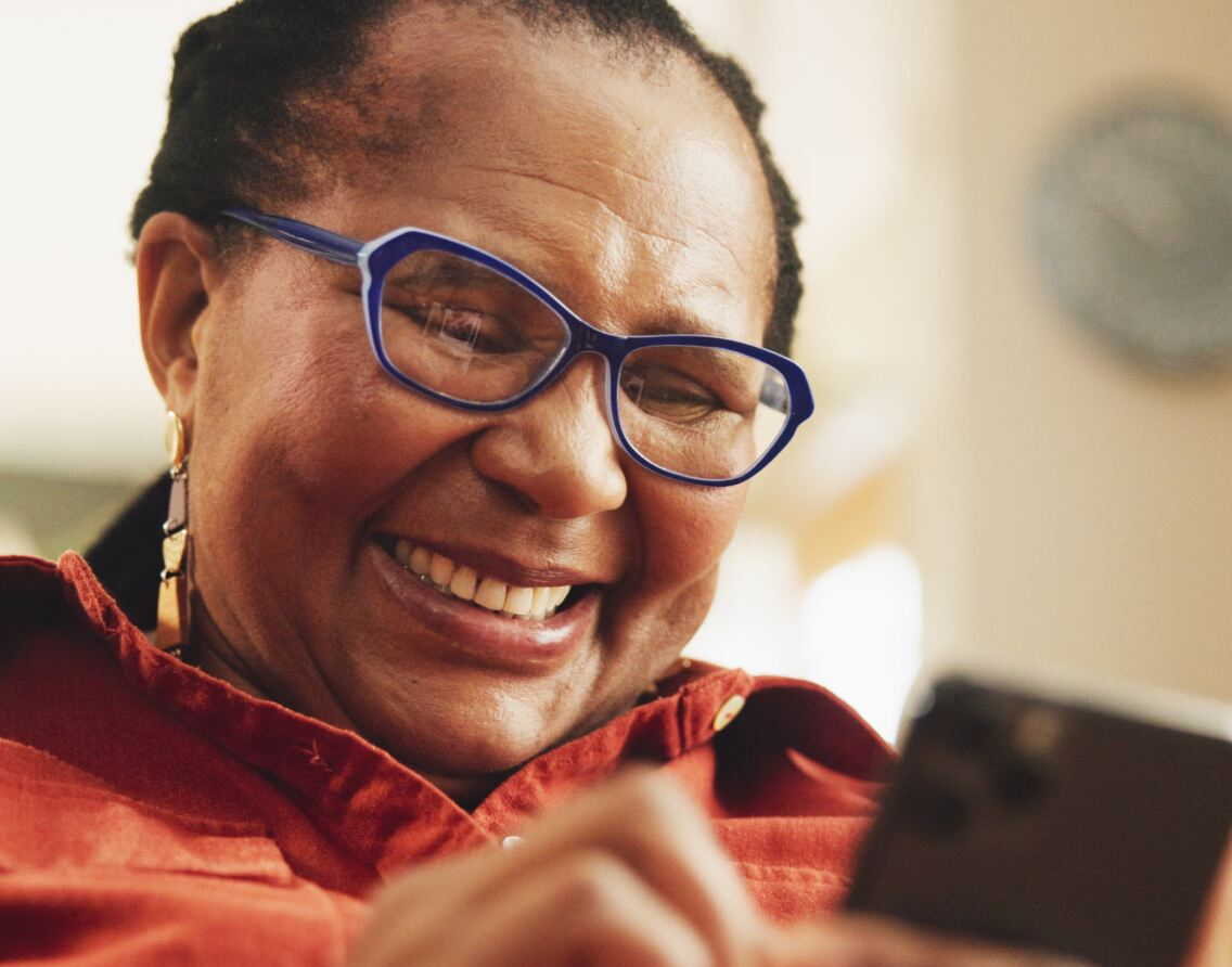 A smiling woman wearing blue glasses and earrings holds and interacts with a smartphone. She is in a warmly lit indoor space with blurred background details, including a round wall clock. No visible text.