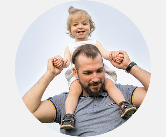 A man wearing a gray polo shirt holds a smiling toddler on his shoulders outdoors under a bright sky. The toddler, dressed in a white outfit, grips the man’s hands.