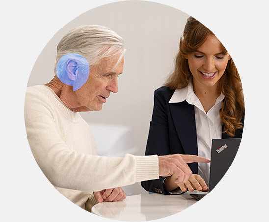 A man consults with a female audiologist in a hearing clinic, pointing at a laptop. His ear is highlighted in blue. Context suggests discussion about hearing aids or tests.