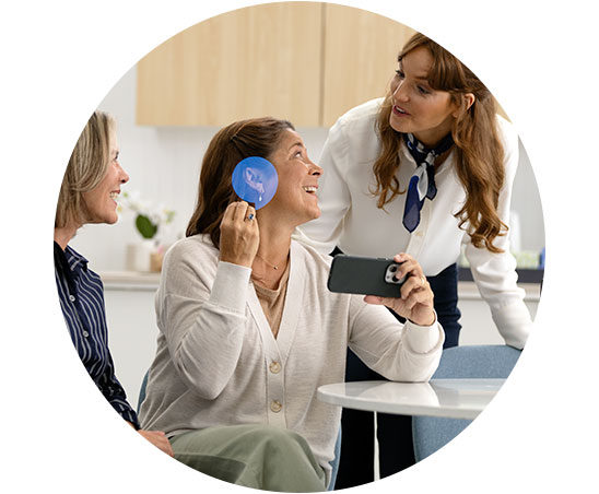 A woman holds a phone and a blue circular tool near her ear, likely in a hearing clinic, as another woman, possibly an audiologist, leans over to assist. The setting includes chairs, a table, and a modern background with a cabinet. No visible text or identifiable hearing aid brands are present.