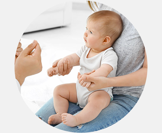 A baby, seated on an adult's lap, attentively looks at another person gesturing with their hands. The setting is a bright indoor space featuring soft furnishings and casual attire.