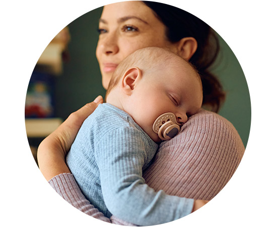 A serene infant sleeps peacefully with a pacifier while being lovingly held by a caregiver in a cozy indoor environment. The background features blurred household items, enhancing the warmth.