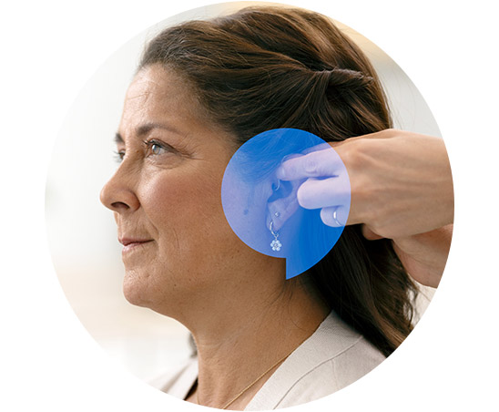 A woman receives a hearing aid adjustment from a professional in a hearing clinic. The setting is bright, with a focus on the ear and hand interaction.