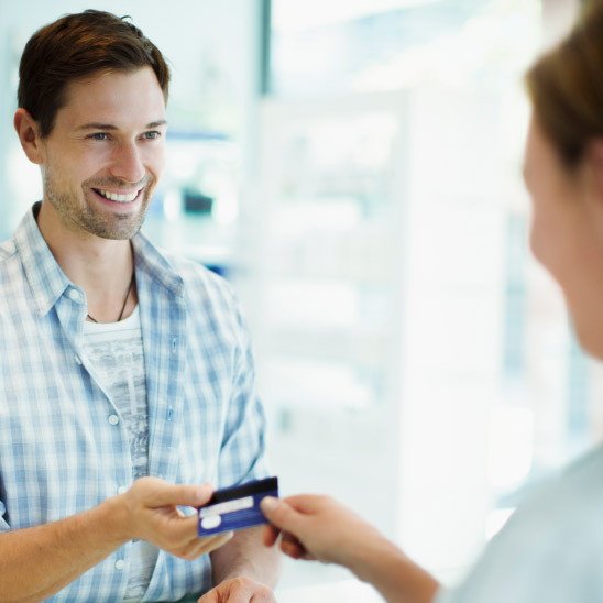 A man in a checkered shirt hands over a credit card to a person, set in a bright, modern hearing clinic reception area. Shelves and light-colored fixtures are visible in the background.