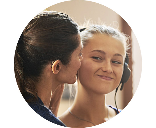 Two people lean closely together; one wears a hearing aid, the other wears headphones and smiles softly, with a gently blurred indoor background and warm lighting.