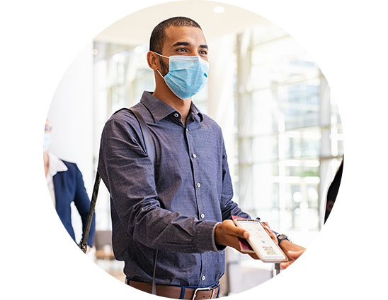A man in a blue shirt and face mask offers a smartphone to another person in a brightly lit, modern indoor space with large windows.