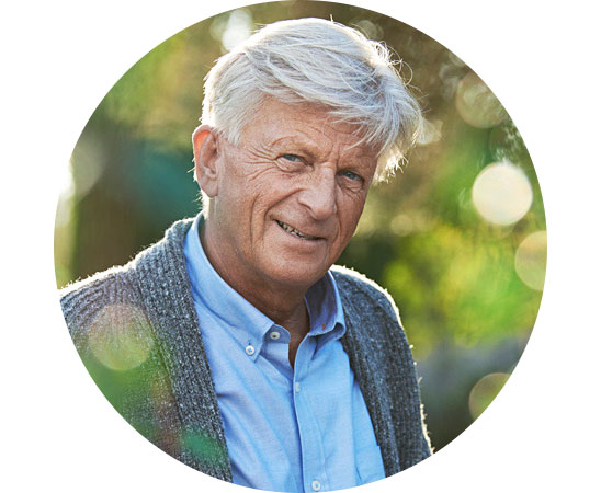 Elderly man smiling outdoors with sunlight and greenery in the background, wearing a blue shirt and gray sweater.
