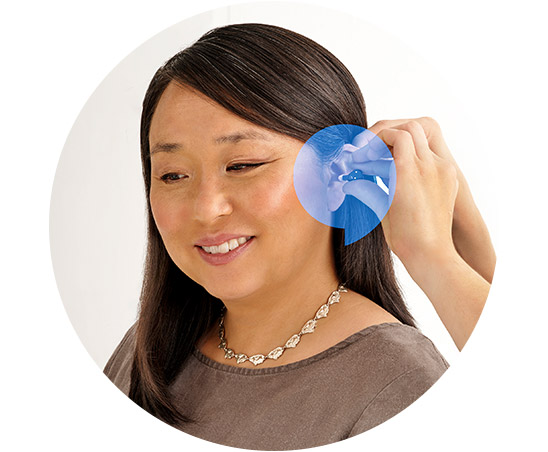 A woman smiles as an audiologist adjusts her small, discreet hearing aid in a professional hearing clinic setting. A circular overlay magnifies the device placement behind her ear for detail.