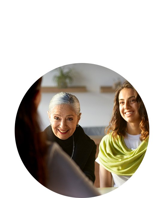 A small group of people sit at a table in a bright, modern room with shelves and plants visible in the background, engaging in a focused discussion. No visible text or distinguishable hearing aid brands appear.