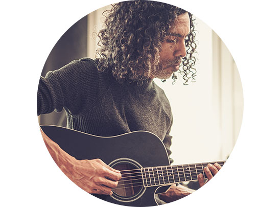 A person with curly dark hair strums an acoustic guitar, seated indoors near softly lit windows, wearing a dark sweater. The focus highlights music and creativity in a serene setting. No visible text or hearing aids related to HearingLife, Oticon, Bernafon, or Philips.