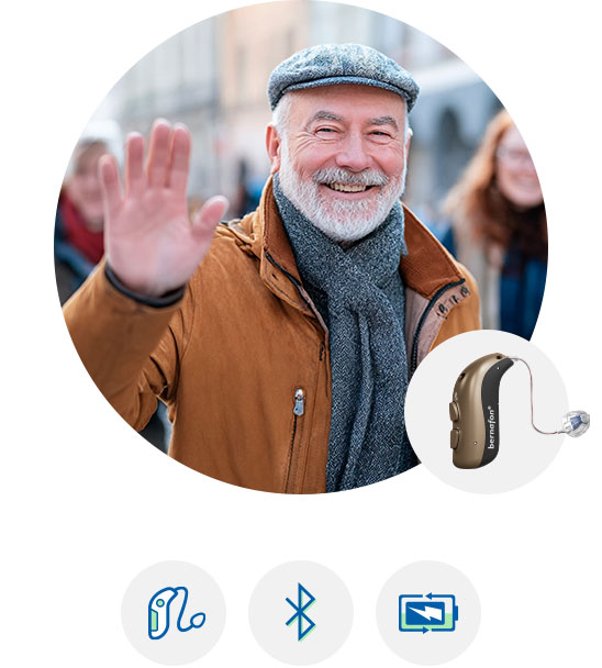 Woman with short gray hair wearing a blue blouse, looking at herself in the mirror next to a close-up of a modern hearing aid device.