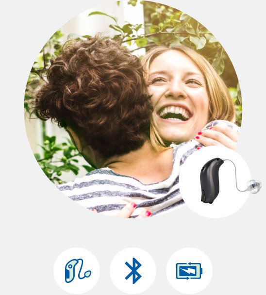 Two women embracing outdoors with hearing aid features displayed below, including Bluetooth connectivity and rechargeable battery.