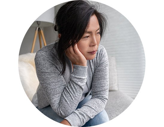 A woman is sitting thoughtfully on a grey couch, resting her head on her hand, in a modern living room with a lamp and a furry cushion behind her.
