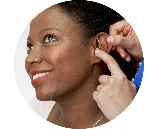 Hearing aid, being placed in a smiling woman's ear, closely framed by two hands, against a neutral background.