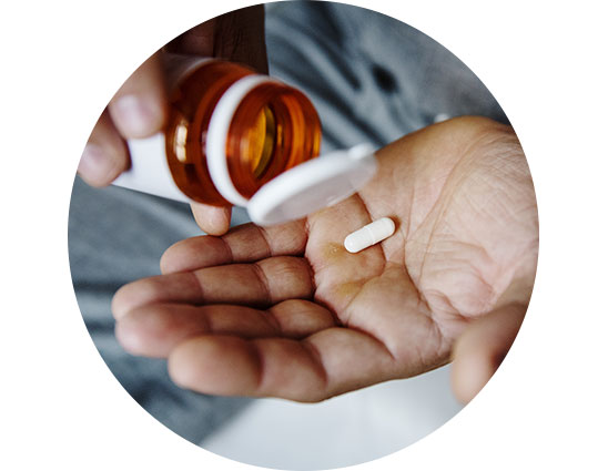 A hand pours a white pill from an orange prescription bottle into the palm, suggesting medication use. The background appears blurred, emphasizing health and wellness in a personal setting. No text visible.