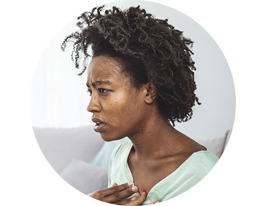A person with short, curly hair sits indoors on a couch, wearing a light green shirt and touching their chest with one hand. No visible hearing aids or text are present.
