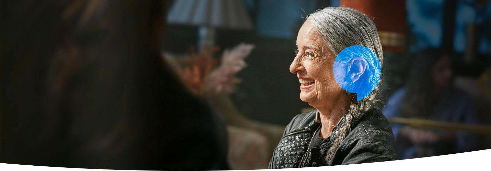 Une femme en veste de cuir est assise dans un intérieur agréable. Une bulle bleue sur son oreille démontre l’importance des appareils auditifs en soins auditifs.