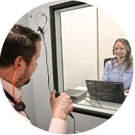 Un patient dans une cabine insonorisée manipule un appareil de test auditif, observé à travers une vitre par une audiologiste avec un portable.